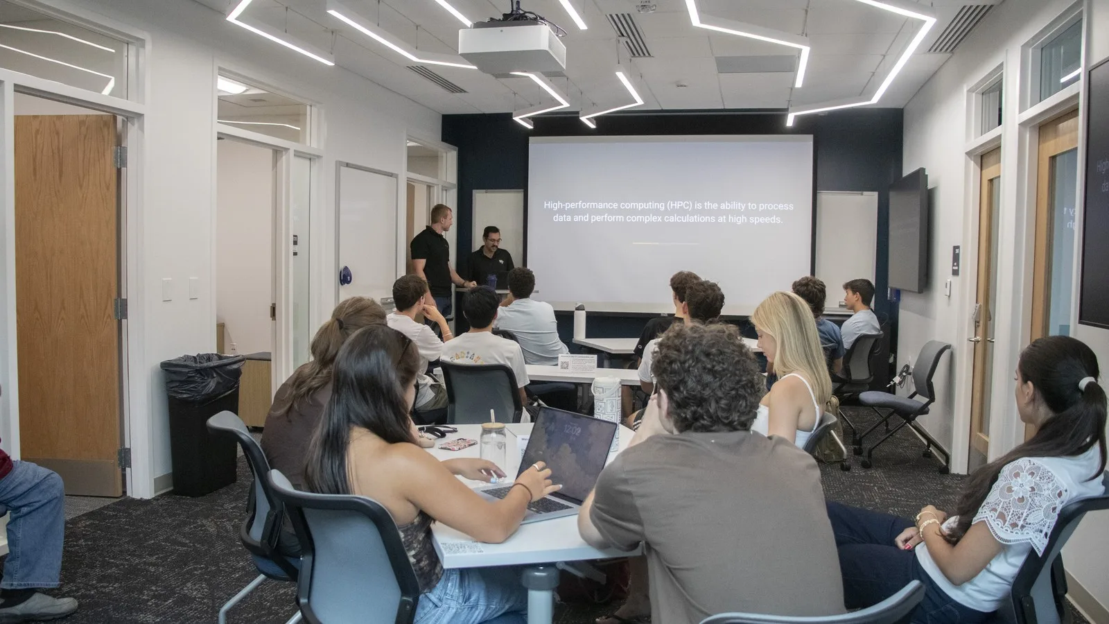Wide classroom view of FYS 100 students seated at tables while two presenters introduce high-performance computing on a projected slide.